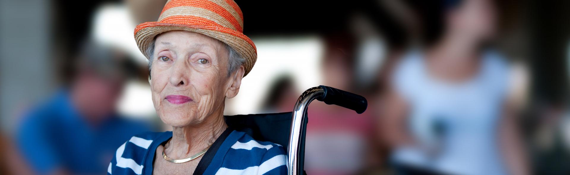 Older white woman wearing an orange striped fedora and blue and white striped shirt sitting in a wheelchair.