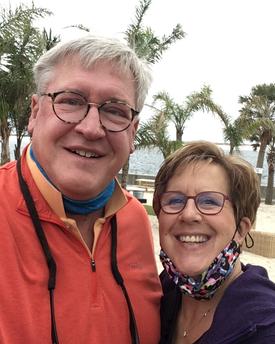 A man and woman are smiling at the camera while standing on a beach with palm trees and a body of water in the background. They are wearing jackets and glasses.